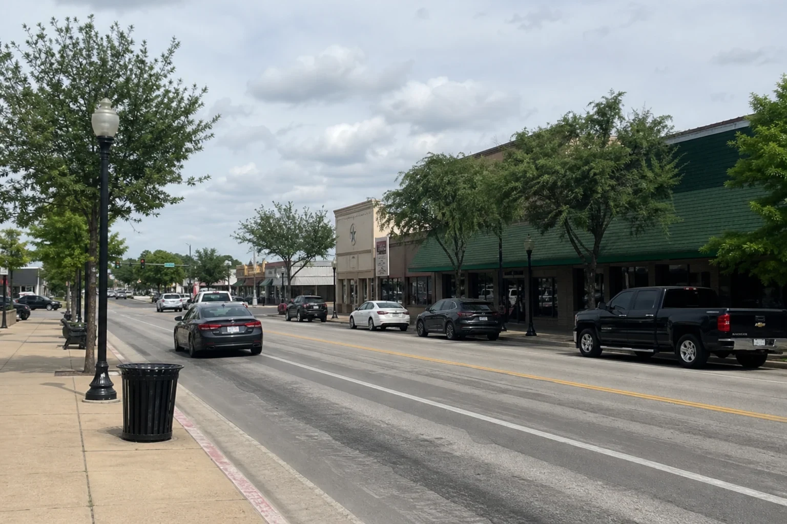 Downtown main street with a wide road, parked cars and a pickup along tree-lined storefronts, street lamps and a trash can on the sidewalk under a cloudy sky